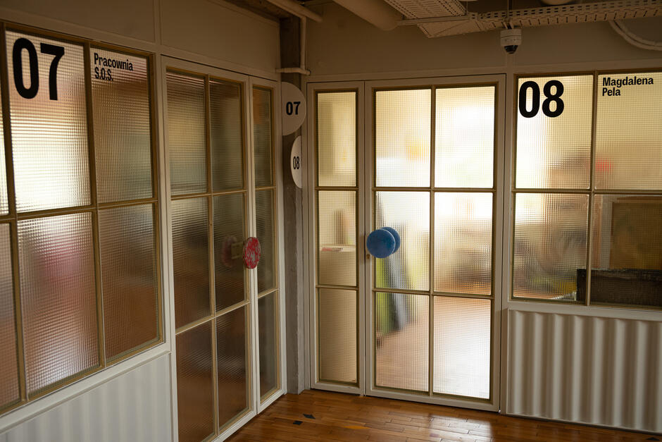 Widać przeszklone drzwi do dwóch pracowni oznaczonych numerami 07 i 08, z nazwiskami właścicieli na szybach. Panuje ciepłe, przytłumione światło, a na klamkach wiszą kolorowe ozdoby przypominające tkaniny.