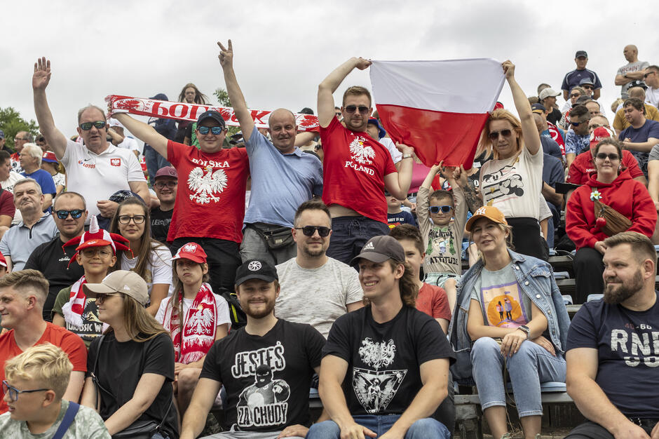 na zdjęciu trybuny wypełnione dziesiątkami kibiców, niektórzy trzymają biało czerwone szaliki i flagi