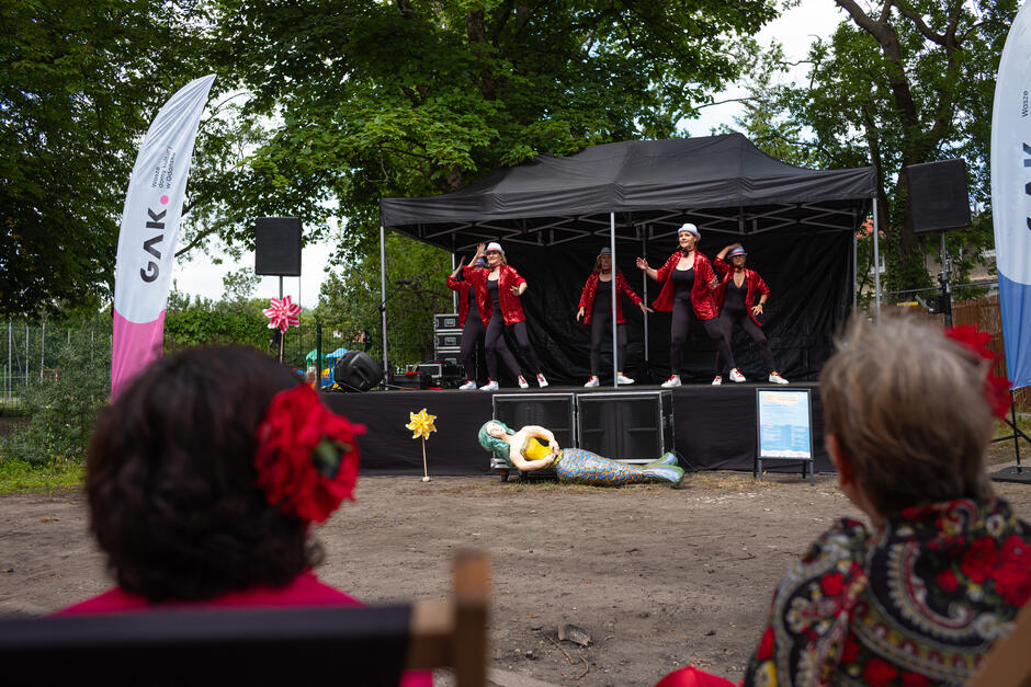 na pierwszym planie kilka osób siedzi, przed nimi scena na której występują kobiety w tanecznych sukniach