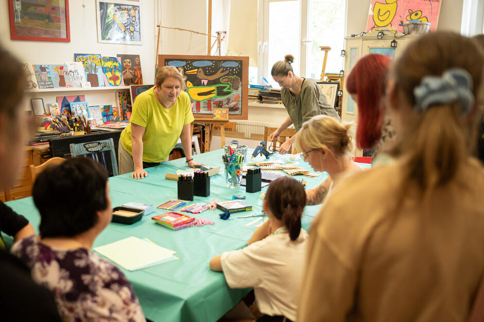 na zdjęciu niewielkie pomieszczenie, na ścianach widać obrazy, na środku stoi duży stół przy którym siedzi kilka dorosłych osób i dzieci, niektóre malują na kartkach