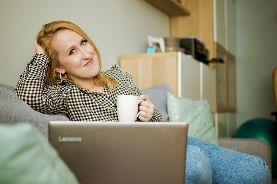 Na zdjęciu kobieta siedząca na kanapie, trzymająca w lewej ręce biały kubek. Przed nią monitor laptopa