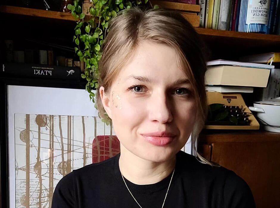 Na zdjęciu kobieta o blond włosach w czarnej bluzce, w tle regał z książkami i roślinami.