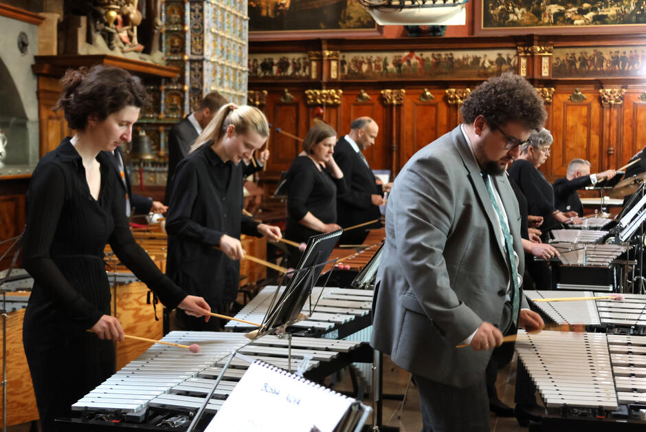 Muzycy Orkiestry Vita Activa grają na instrumentach perkusyjnych melodycznych, takich jak wibrafony i dzwonki, z pełnym skupieniem i zaangażowaniem. W tle widać bogato zdobione wnętrze historycznego Dworu Artusa oraz innych członków zespołu