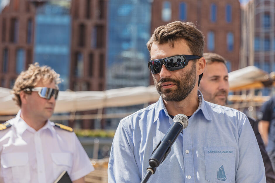 Na zdjęciu widzimy mężczyznę w niebieskiej koszuli z logo „Generał Zaruski”, który przemawia do mikrofonu podczas wydarzenia plenerowego, najprawdopodobniej związanego z żeglarstwem. W tle znajdują się inni uczestnicy, a także nowoczesne budynki i fragment żaglowca.