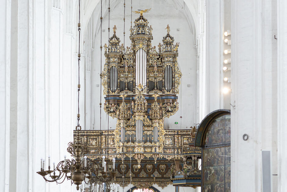 Na zdjęciu widać bogato zdobione, monumentalne organy w Bazylice Mariackiej w Gdańsku, wyróżniające się złoceniami, rzeźbami i kunsztowną ornamentyką. Wnętrze świątyni jest jasne i przestronne, z wysokimi, białymi ścianami oraz widocznymi żyrandolami i elementami ołtarza