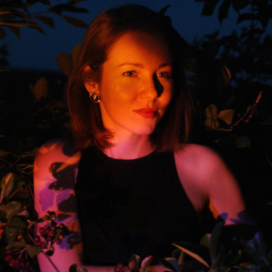 Ma zdjęciu kobieta w czarnej koszulce, z gołymi ramionami, otulona w liście drzewa.