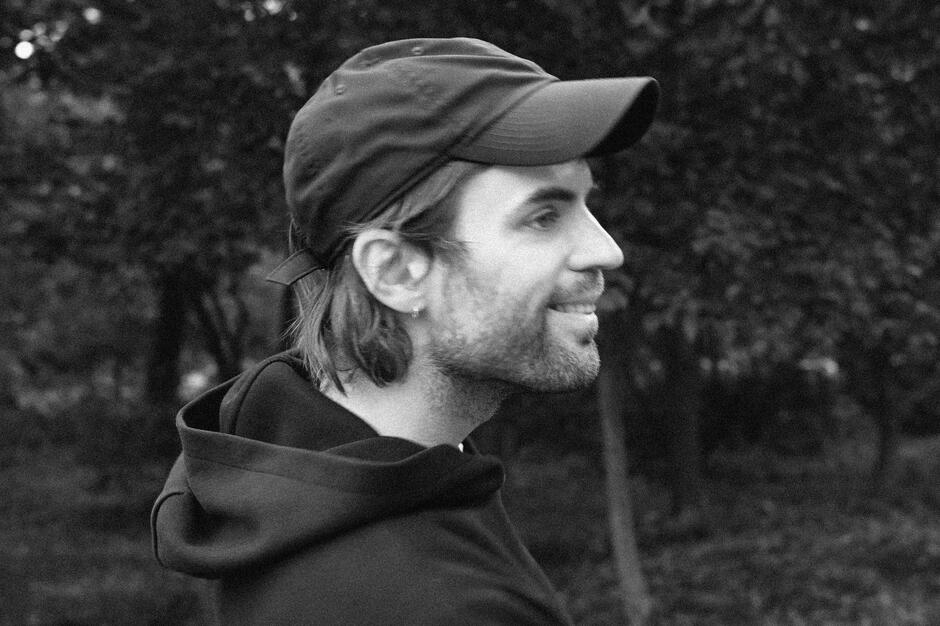 Na zdjęciu mężczyzna patrzący w prawo, na sobie ma bluzę z czarnym kapturem i czapeczkę z daszkiem. Na twarzy lekki zarost.