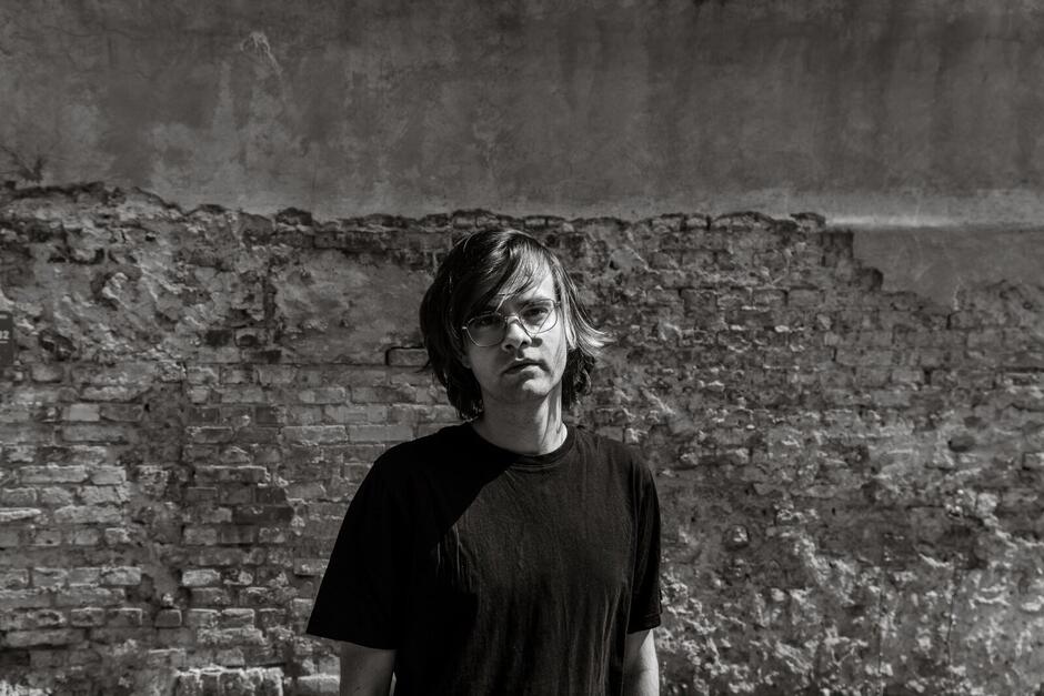 Na zdjęciu mężczyzna z włosami za uszy, ubrany w czarny T-shirt, na tle zniszczonej ściany.