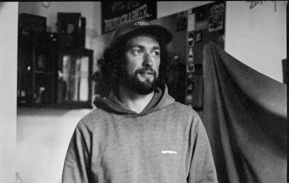 Na zdjęciu mężczyzna z włosami za uszy, z zarostem, na głowie ma czapkę z daszkiem, ubrany w bluzę. 