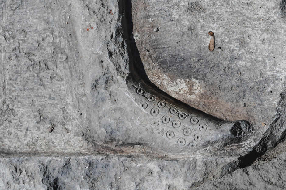Zbliżenie na detal kamiennego reliefu, gdzie wyraźnie widać nogę w średniowiecznym bucie z ozdobnym wzorem w postaci małych, regularnych kręgów, przypominających łuski lub dekoracyjne tłoczenia