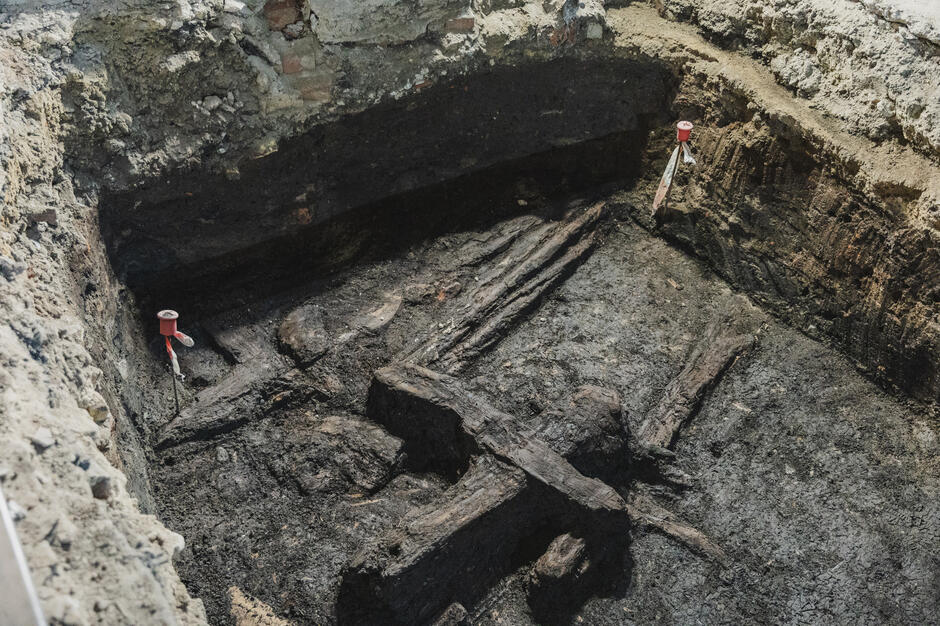 Ukazuje część wykopu archeologicznego z dobrze zachowanymi fragmentami drewnianych konstrukcji, prawdopodobnie dawnych fundamentów lub umocnień. Ziemia jest ciemna i wilgotna, co sugeruje długoletnie zaleganie drewna w warunkach ograniczonego dostępu powietrza.