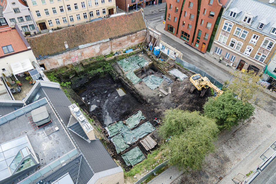 Widok z lotu ptaka na całe stanowisko archeologiczne w środku miasta, gdzie widać kilka odkrytych kwater wykopaliskowych przykrytych plandekami oraz ciężki sprzęt budowlany. Całość otoczona jest współczesną zabudową miejską, co pokazuje kontrast między dawną historią a nowoczesnością.