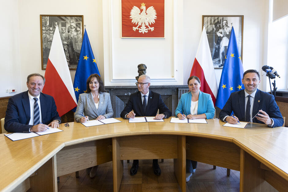 Na zdjęciu widzimy pięć osób siedzących przy dużym, drewnianym stole, podpisujących dokumenty. W tle widoczne są flagi Polski i Unii Europejskiej oraz godło Polski (biały orzeł na czerwonym tle). Za osobami stoją także dwa czarno-białe portrety historycznych postaci w wojskowych mundurach. Centralnie na ścianie umieszczone jest popiersie. Osoby na zdjęciu ubrane są formalnie – mężczyźni w garniturach i kobiety w eleganckich żakietach. Sytuacja wygląda na oficjalne podpisanie umowy lub listu intencyjnego, prawdopodobnie dotyczącego współpracy między instytucjami publicznymi lub samorządowymi w Polsce. Atmosfera jest uroczysta i oficjalna, co dodatkowo podkreślają symbole narodowe i unijne obecne w pomieszczeniu.