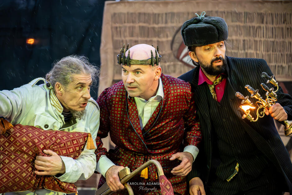 Trzej aktorzy w kostiumach teatralnych w napięciu pochylają się ku widzowi, każdy z wyrazistą mimiką i rekwizytem. Atmosfera zdjęcia jest pełna dramatyzmu i skupienia