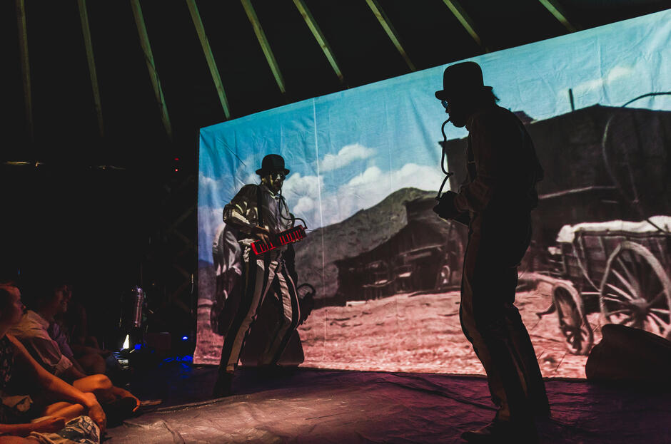 Dwóch performerów na tle projekcji filmowej nawiązuje do estetyki niemego kina, z teatralnymi gestami i stylizacją retro. Kontrast między światłem projekcji a ciemnością sali buduje efekt teatralnego spektaklu wizualnego