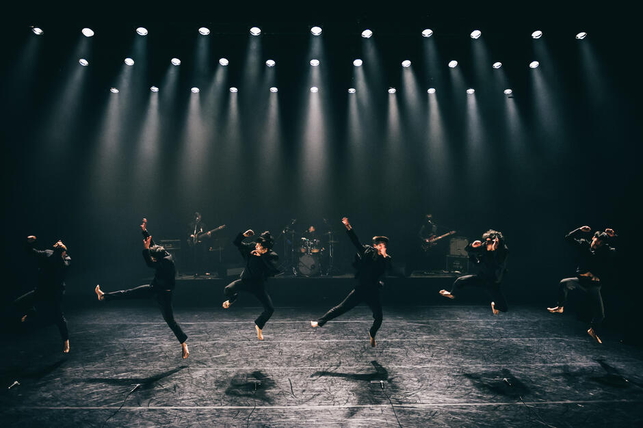 Grupa tancerzy wykonuje synchroniczny skok na scenie w rytm muzyki na żywo grającego zespołu. Minimalistyczne oświetlenie i ciemne stroje tworzą dynamiczny i ekspresyjny obraz