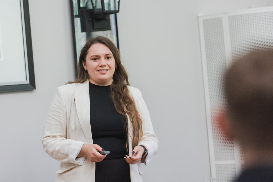 Na zdjęciu kobieta w jasnej marynarce i czarnej bluzce prowadzi prezentację, trzymając w rękach pilot do slajdów. Uśmiechnięta prelegentka stoi przed grupą słuchaczy, z których część jest widoczna na pierwszym planie. 