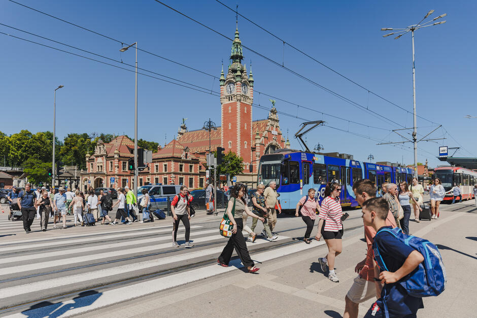 Na zdjęciu widać grupę pieszych przechodzących przez szerokie przejście dla pieszych w centrum Gdańska, na tle charakterystycznego budynku Dworca Głównego PKP. Obok zatrzymał się niebieski tramwaj, a nad ulicą przebiegają liczne linie trakcyjne pod jasnym, bezchmurnym niebem. 
