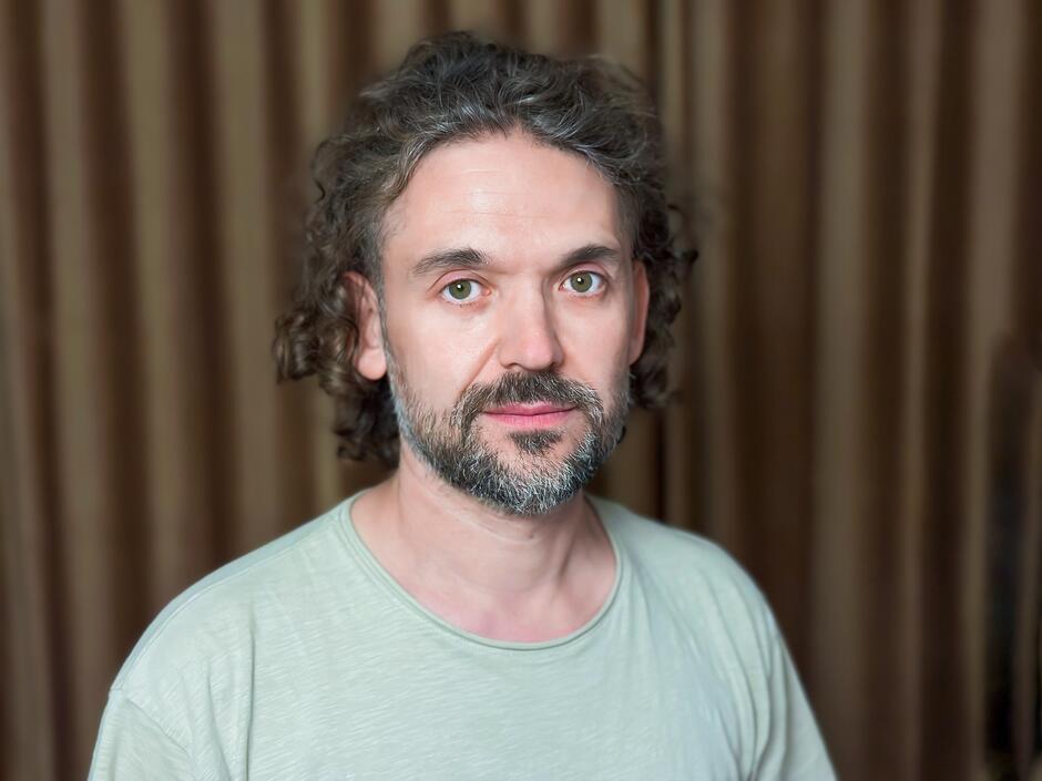 Na zdjęciu mężczyzna z kręconymi włosami i zarostem. Na tle brązowej kotary.