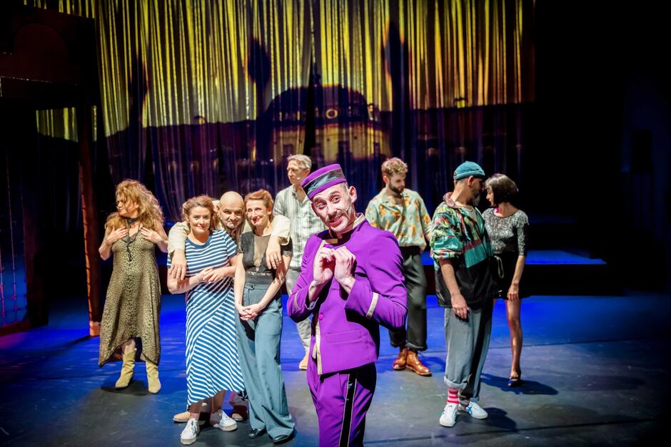 Na zdjęciu widzimy grupę aktorów na scenie teatralnej, z których jeden – ubrany w charakterystyczny fioletowy kostium przypominający strój boy’a hotelowego – wyraźnie pozuje na pierwszy plan z zabawną miną. W tle reszta zespołu znajduje się w różnych pozach, a całość dopełnia kolorowa, teatralna scenografia z podświetlonym tłem przypominającym wnętrze luksusowego hotelu