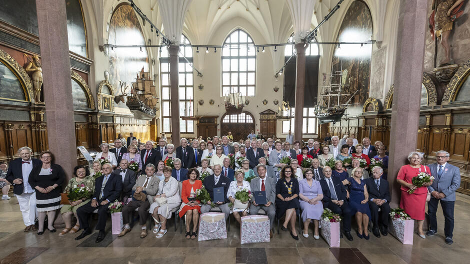 Zdjęcie przedstawia dużą grupę starszych osób zgromadzonych w zabytkowej sali z wysokimi sklepieniami, ozdobionej modelami żaglowców i malowidłami ściennymi – prawdopodobnie podczas uroczystości jubileuszu małżeńskiego, tzw. "Złotych Godów". Wiele par trzyma bukiety kwiatów i dyplomy, a atmosfera jest odświętna i pełna radości