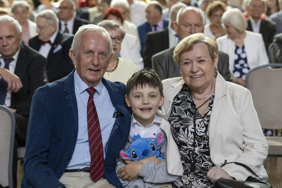 Na zdjęciu widać starsze małżeństwo siedzące wśród innych uczestników uroczystości, uśmiechających się do kamery wraz z wnuczkiem, który trzyma w ramionach maskotkę Stitcha. W tle znajduje się liczna grupa elegancko ubranych seniorów, co sugeruje, że zdjęcie wykonano podczas oficjalnego wydarzenia, prawdopodobnie jubileuszu złotych godów