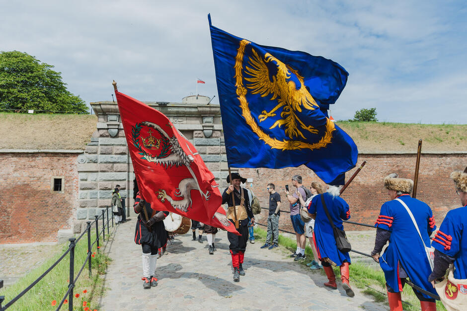 Dwaj mężczyźni z powiewającymi flagami, wokół nich kilku innych mężczyzn - wszyscy w historycznych strojach z XVII wieku
