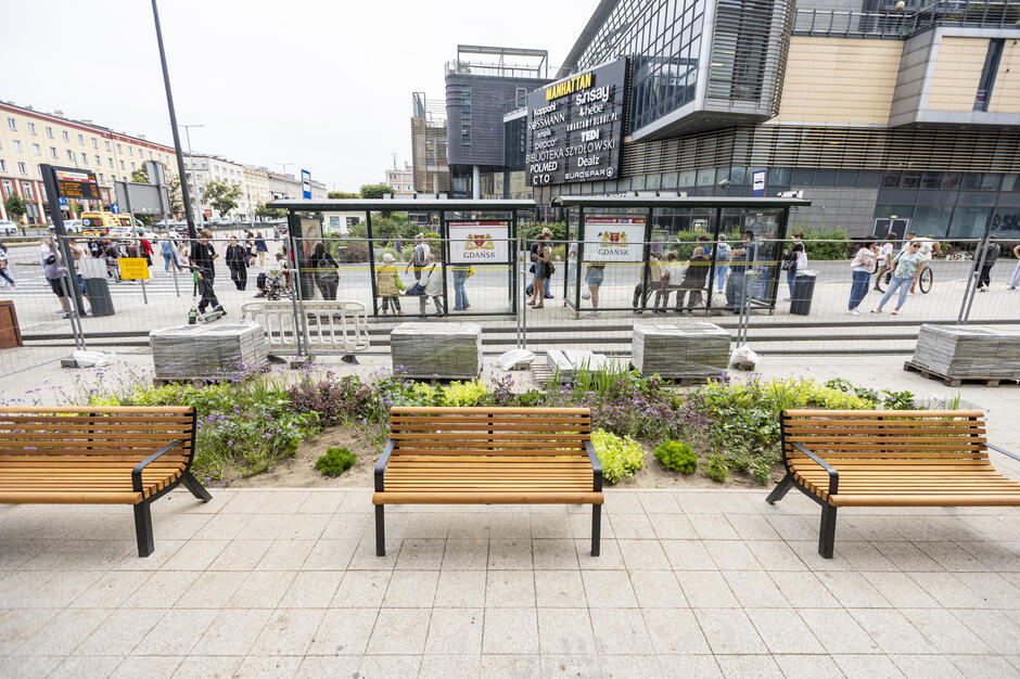 Zdjęcie ukazuje odnowioną przestrzeń miejską przed centrum handlowym Manhattan w Gdańsku, z nowymi ławkami i kolorową rabatą kwiatową. W tle widoczny jest przystanek tramwajowy z licznymi pasażerami oraz charakterystyczny budynek centrum handlowego.