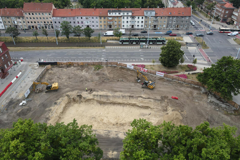 zdjęcie z drona, na dole zdjęcia plac budowy z koparkami i ciężarówkami, wyżej jezdnia trzypasmowa którą jadą samochody, w tle niewysokie ceglane kamienice