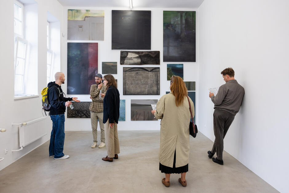 Zdjęcie przedstawia grupę osób oglądających współczesną wystawę sztuki w jasnej, minimalistycznej galerii z wieloma abstrakcyjnymi obrazami zawieszonymi na jednej ścianie. Część osób rozmawia ze sobą, a inni indywidualnie analizują dzieła, trzymając w rękach broszury wystawy