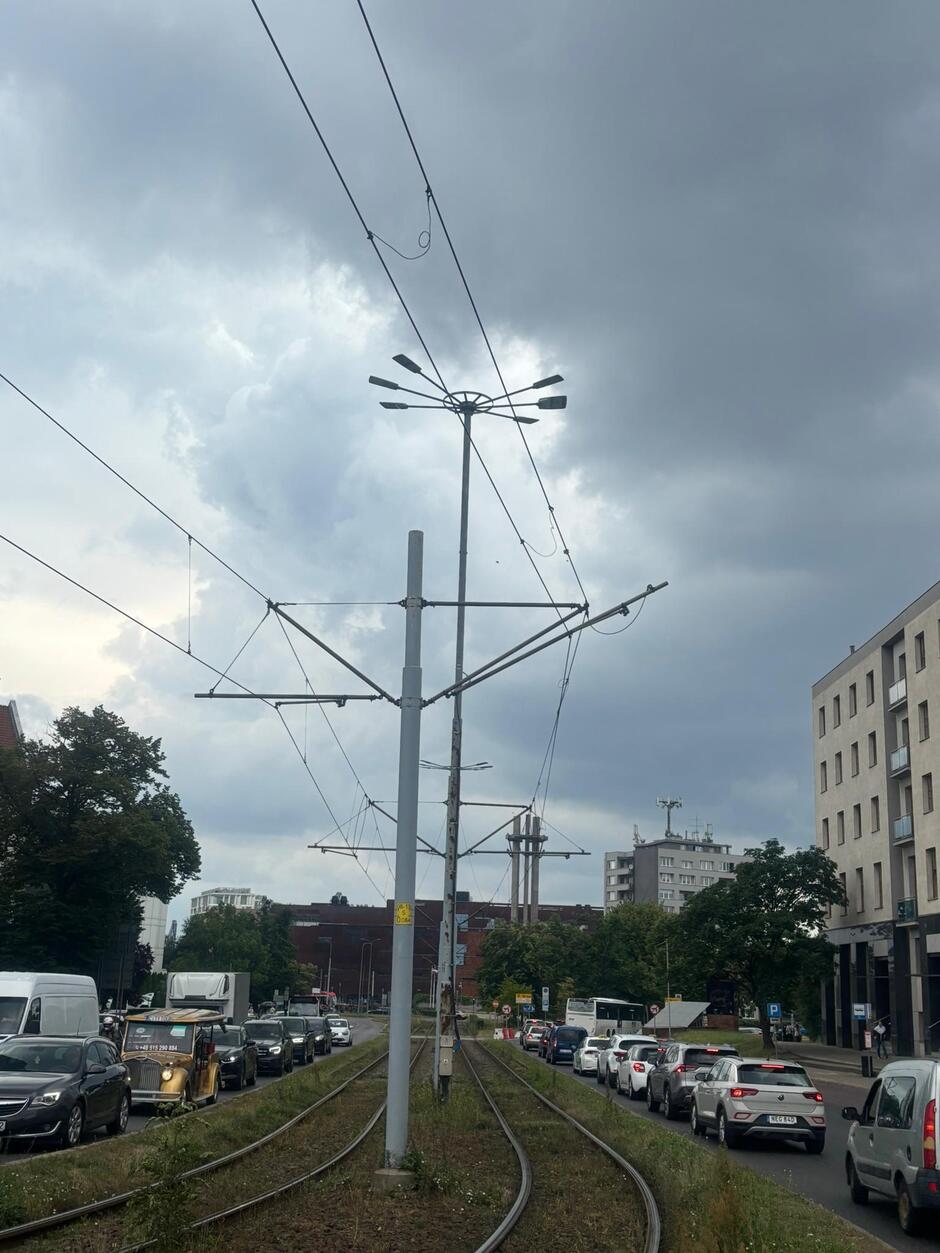 Widok słupów trakcji tramwajowych i torowiska. Po obu stronach torowiska pasy jezdni ze stojącymi na nich samochodami