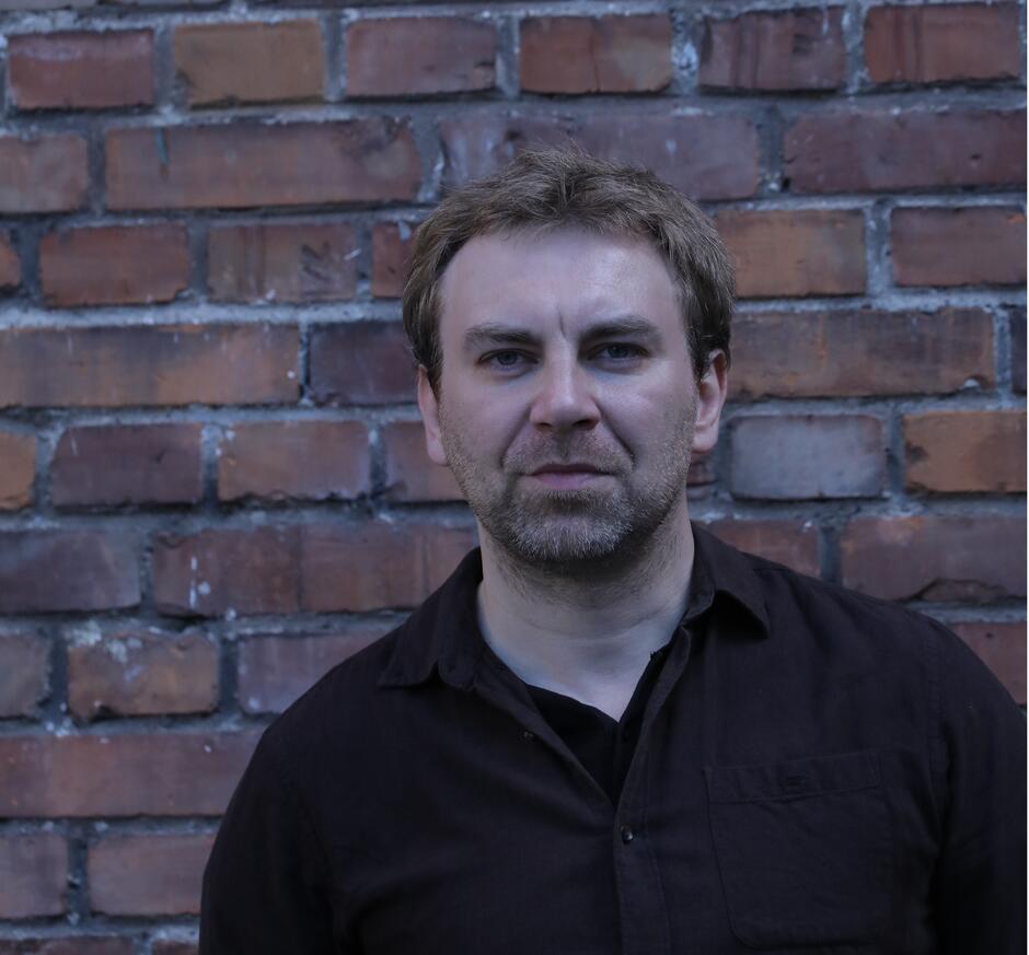 Na zdjęciu mężczyzna z krótkimi włosami i lekkim zarostem na tle ceglanej ściany.