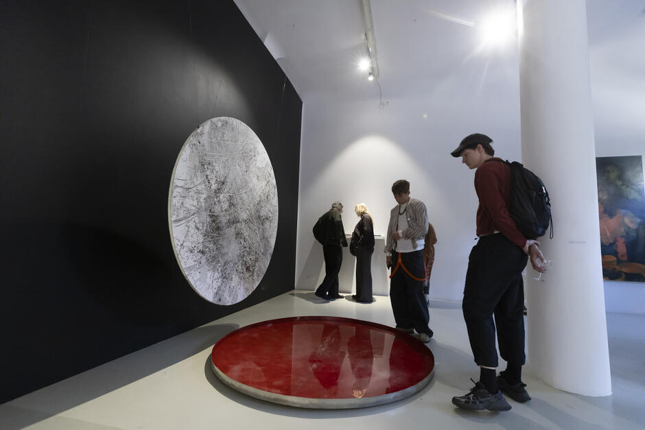 Na zdjęciu widać grupę osób oglądających nowoczesną instalację artystyczną w galerii – na czarnej ścianie zawieszony jest okrągły obiekt przypominający zarysowany księżyc, a poniżej na podłodze znajduje się czerwony, lustrzany krąg. Zwiedzający uważnie przyglądają się dziełu, a atmosfera sprzyja refleksji i kontemplacji sztuki współczesnej.