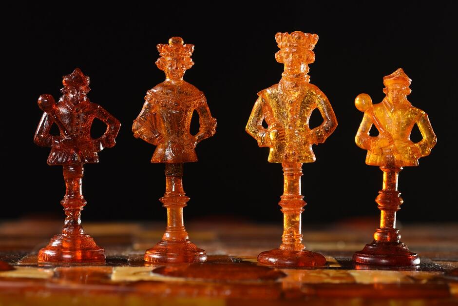 Na zdjęciu widoczne są misternie wykonane bursztynowe figurki szachowe z XVII wieku, przedstawiające postacie w królewskich strojach, każda z charakterystyczną pozą i detalami. Ciepłe światło podkreśla przezroczystość i bogactwo barw bursztynu, tworząc wrażenie luksusu i historycznego kunsztu.