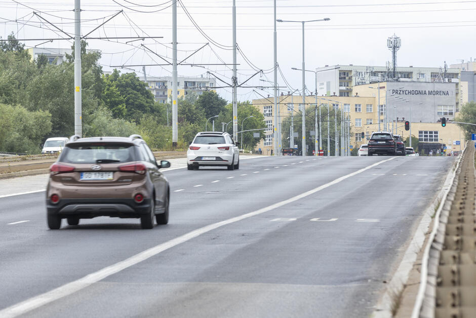 na zdjęciu ulica trzypasmowa, jedzie nią kilka samochodów osobowych, w tle kilka niewysokich budynków