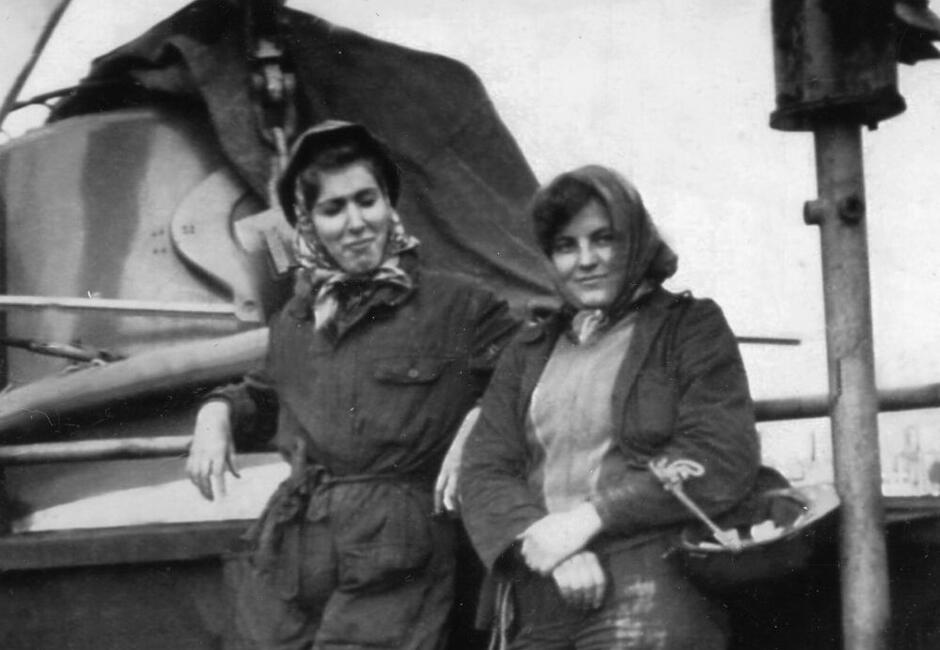 Na czarno-białym zdjęciu widoczne są dwie kobiety ubrane w robocze kombinezony i chustki na głowach, stojące obok siebie na tle przemysłowego sprzętu, prawdopodobnie w stoczni. Ich postawa i wyraz twarzy sugerują dumę i koleżeńską więź, oddając ducha pracy kobiet w przemyśle w okresie powojennym