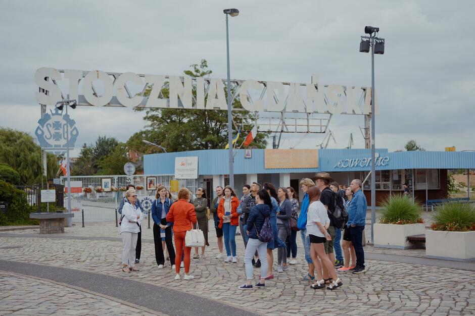Na zdjęciu widać grupę osób biorących udział w oprowadzaniu przed historyczną bramą Stoczni Gdańskiej, która odegrała kluczową rolę w polskich przemianach społeczno-politycznych. Charakterystyczny napis „Stocznia Gdańska” w tle oraz industrialna sceneria nadają miejscu symboliczne znaczenie i przypominają o dziedzictwie Solidarności