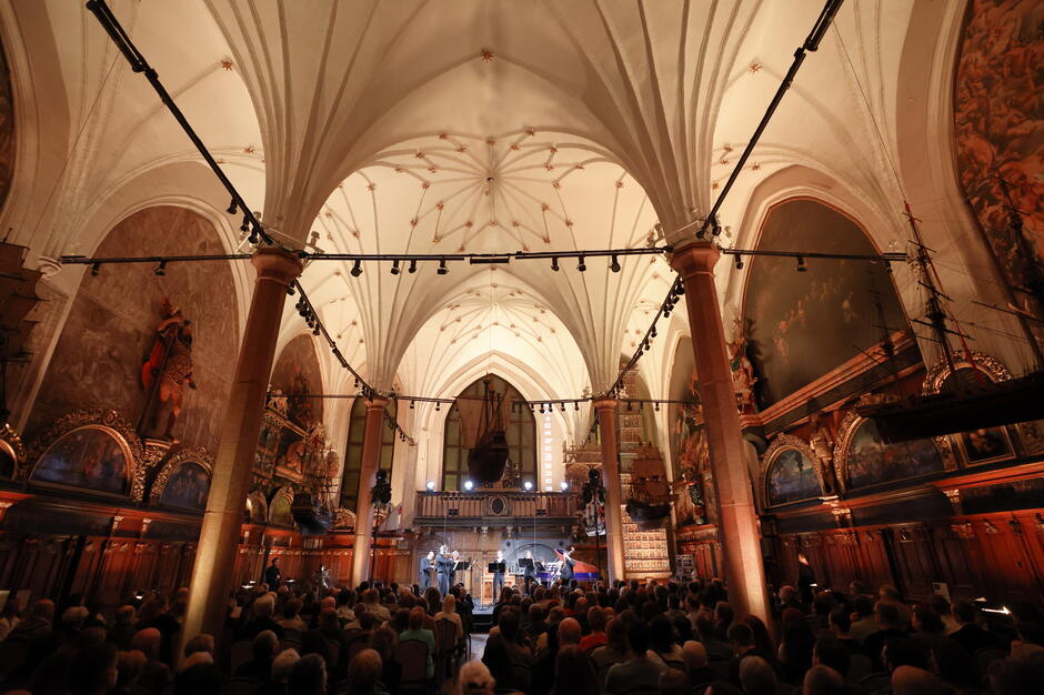 Zdjęcie przedstawia koncert odbywający się w zabytkowym wnętrzu z gotyckim sklepieniem, bogato zdobionymi malowidłami i modelami okrętów zawieszonymi pod sufitem. Przed sceną zgromadzona jest liczna publiczność, wsłuchująca się w występ zespołu muzycznego. 
