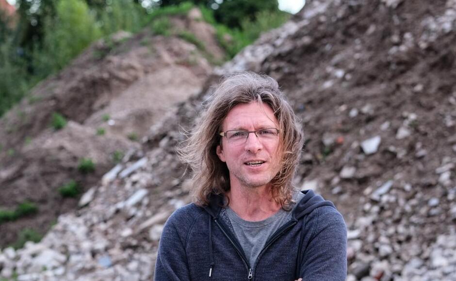 Na zdjęciu mężczyzna z włosami za ramiona, w okularach, na tle nasypów z kamieni i ziemi.