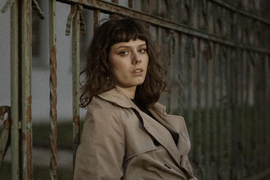 Na zdjęciu kobieta w jasnym prochowcu na tle kutej bramy.