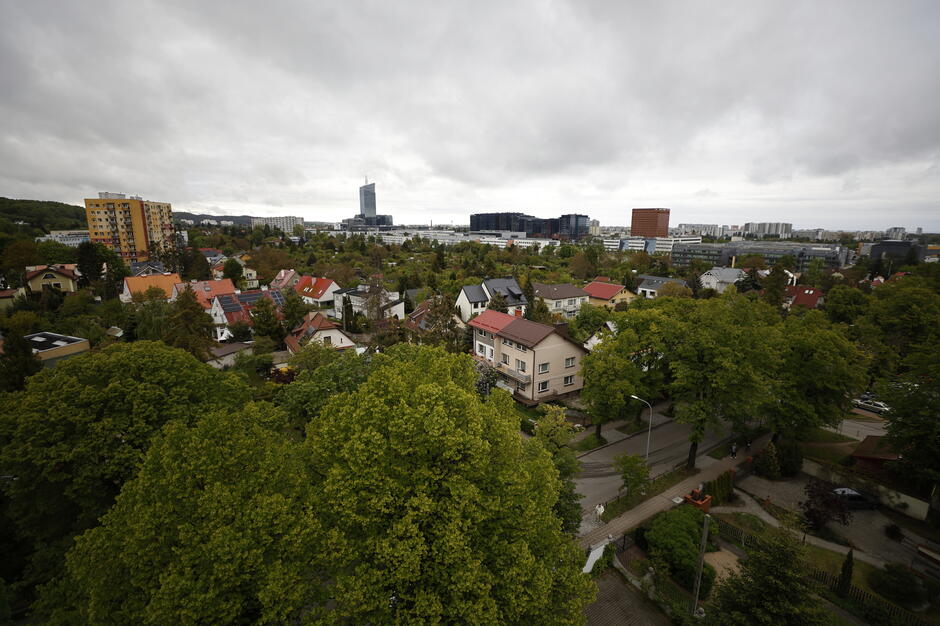 Zdjęcie ukazuje panoramę miejskiej dzielnicy z niską zabudową jednorodzinną, otoczoną gęstą zielenią drzew. W tle widać nowoczesne wieżowce oraz charakterystyczny czerwony budynek, kontrastujące z tradycyjną architekturą osiedla. 