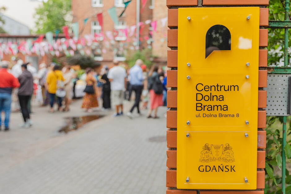 Zdjęcie przedstawia tablicę informacyjną z napisem „Centrum Dolna Brama, ul. Dolna Brama 8, GDAŃSK” umieszczoną na ceglanej kolumnie, przy wejściu do miejskiego ośrodka. W tle widać tłum ludzi biorących udział w wydarzeniu plenerowym, udekorowanym kolorowymi girlandami, co nadaje miejscu atmosferę lokalnego święta lub festynu.
