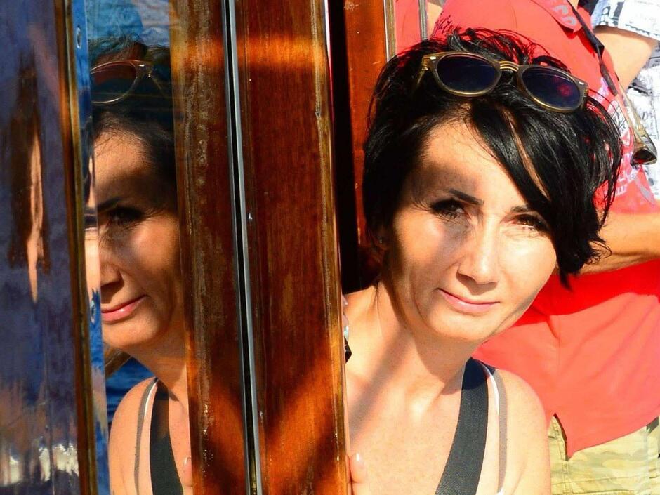 Na zdjęciu kobieta w ciemnych włosach, wyglądająca zza okna.