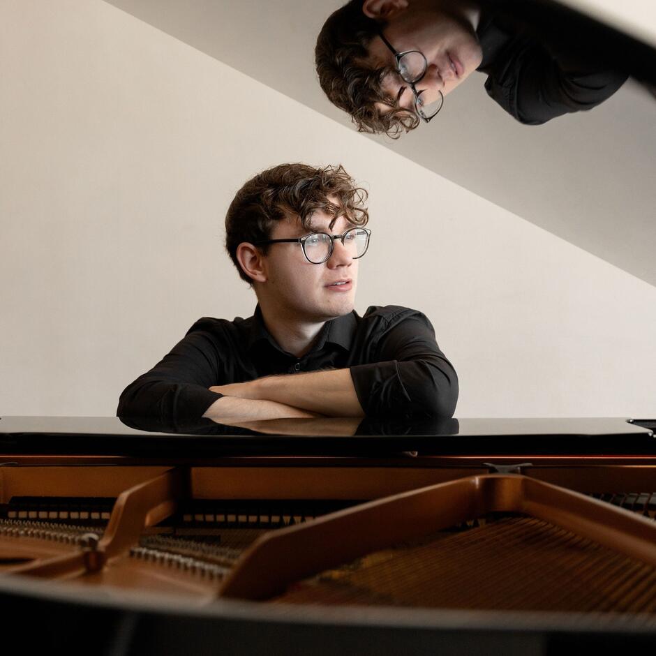Na zdjęciu mężczyzna w okularach oparty o otwartą klapę fortepianu.