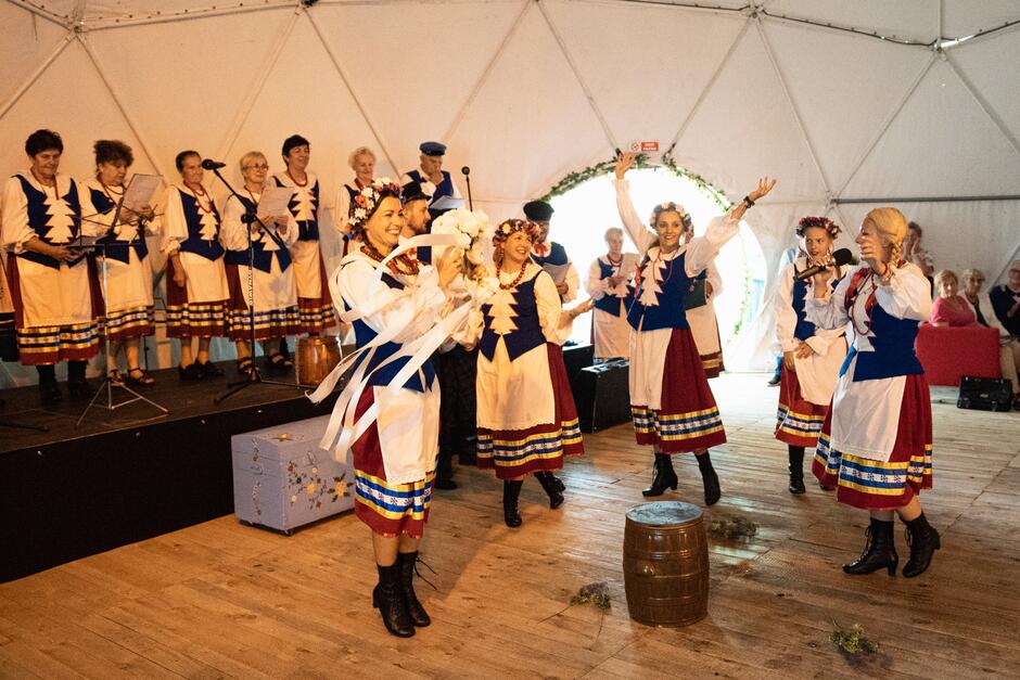 Grupa tancerzy i wokalistów w tradycyjnych strojach kaszubskich występuje na scenie w drewnianym wnętrzu