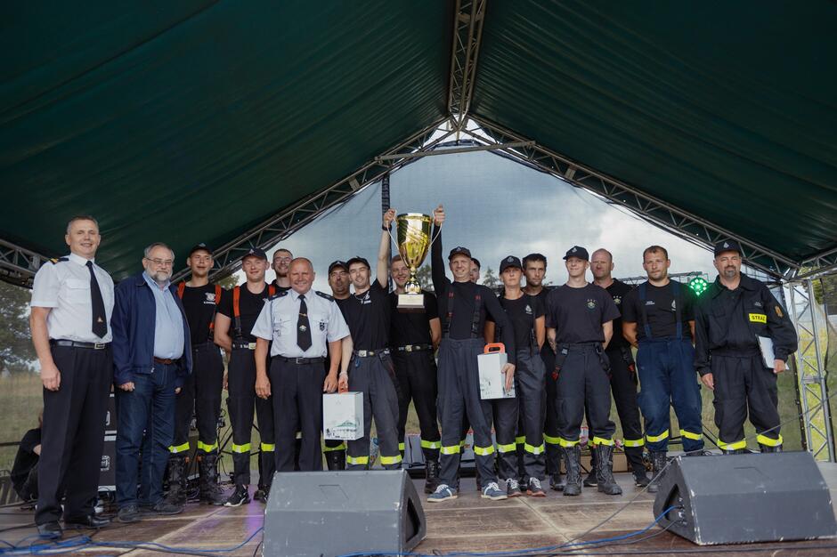 Grupa strażaków pozuje na scenie pod namiotem z dużym pucharem za zwycięstwo w zawodach. Uśmiechy i radość na twarzach świadczą o sportowej atmosferze i udanym wydarzeniu