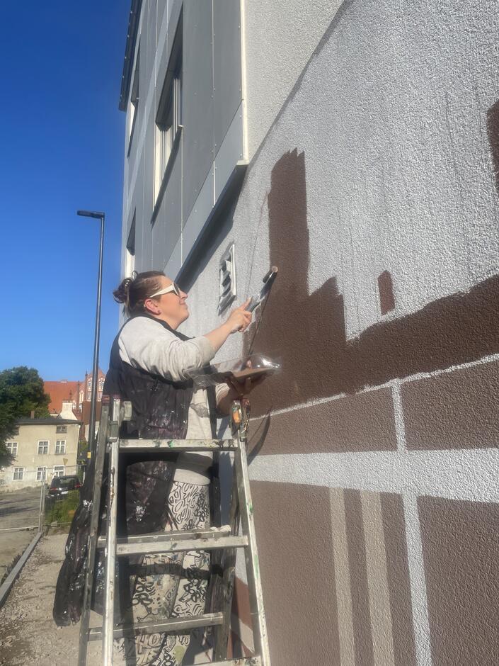 Artystka ostrożnie maluje fasadę budynku wałkiem, tworząc geometryczne kształty w odcieniach brązu.