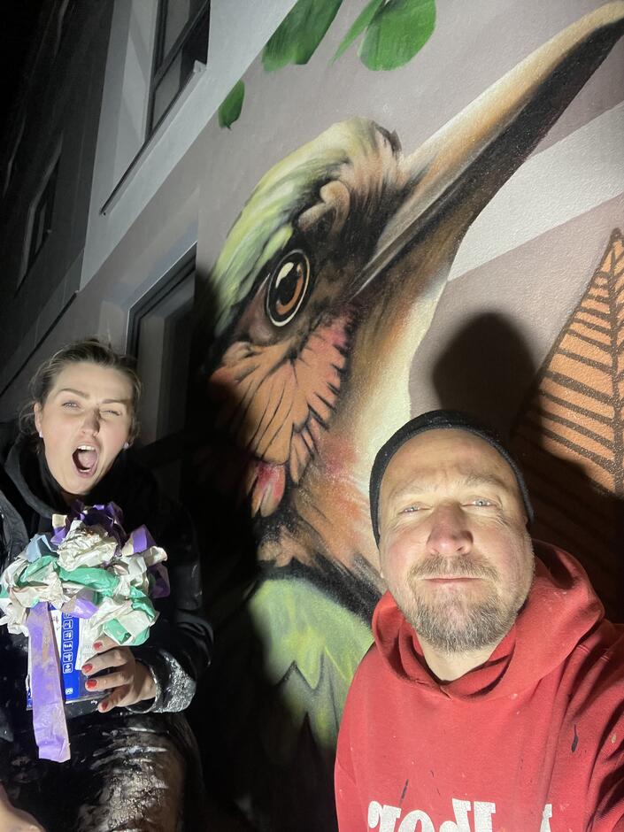 Dwójka twórców robi sobie zdjęcie podczas pracy przy muralu przedstawiającym ptaka, jeden z nich trzyma rolkę popsikaną farbą. Dzięki nocnej perspektywie i światłu latarki scena ma artystyczny klimat, pełen zaangażowania i zabawy