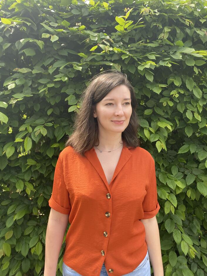Na zdjęciu kobieta z włosami za ramiona, ubrana w pomarańczową letnią bluzkę, na tle zielonych liści.