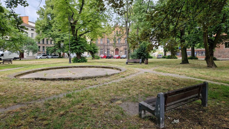 Popękana misa fontanny i zniszczona ławka, wydeptane ścieżki w trawie, z tyłu drzewa a z nimi ceglany zaniedbany budynek 